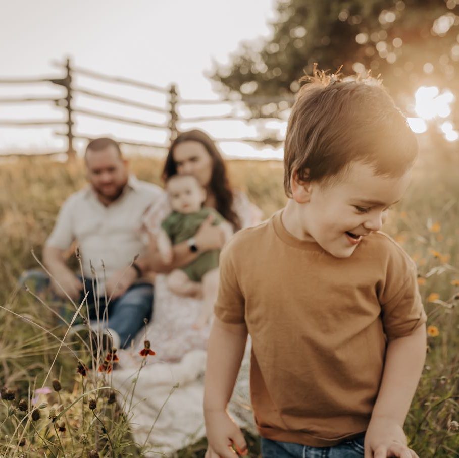 A boy smiling as his parents and siblings sit in the background of a field of tall grass and flowers for a newborn photography session.