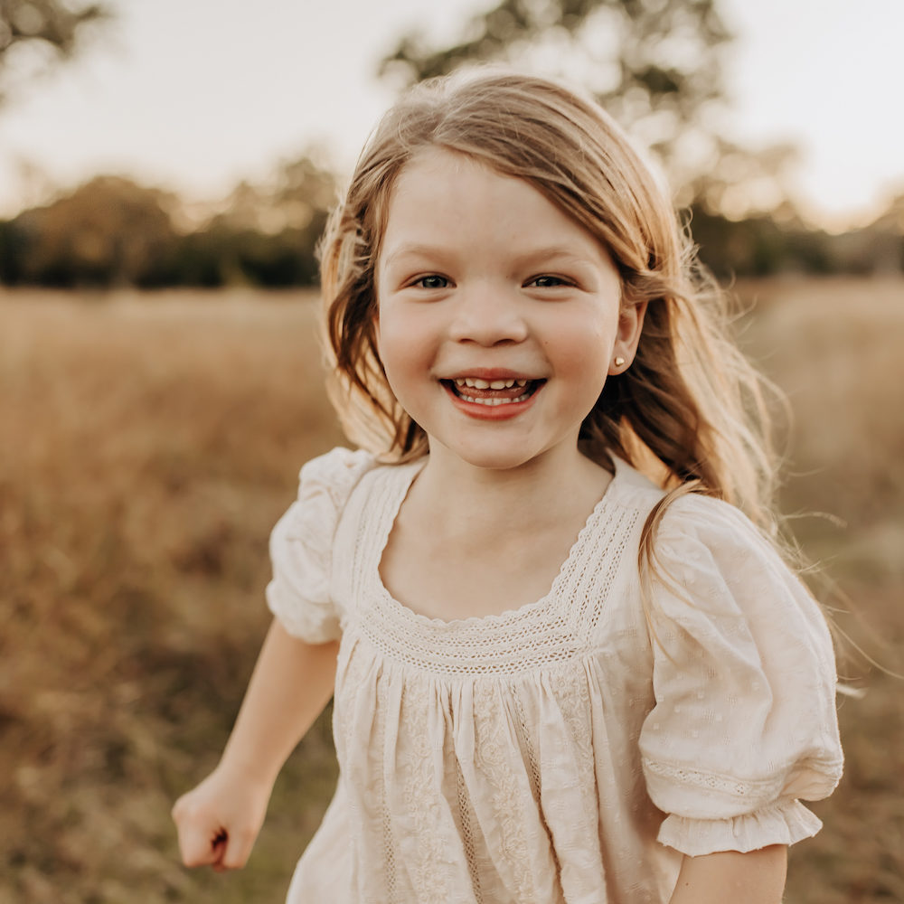 A toddler girl in a cream dress running toward the camera smiling. She is in a San Antonio grassy field during golden hour.